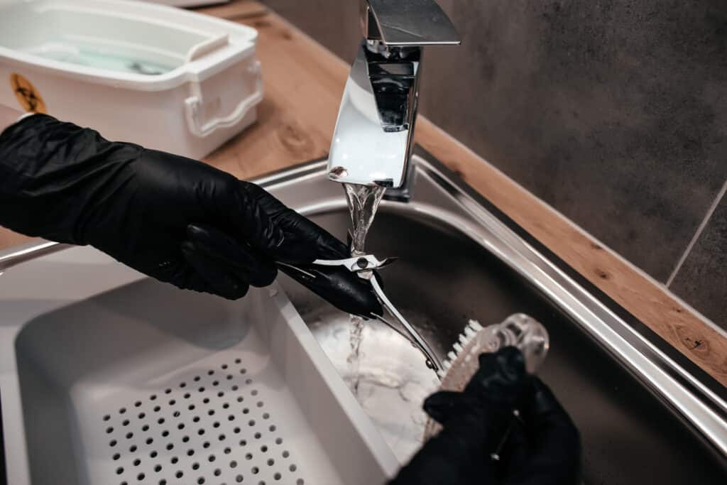 A person wearing black gloves cleaning a pair of nail clippers under running water at a kitchen sink. In the background, a white container can be seen.